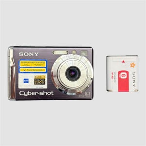 May include: A silver Sony Cyber-shot digital camera with a Carl Zeiss lens and a separate battery. The camera features a Full HD 1080 resolution and 8.1 megapixels. The battery is white with red accents and the Sony logo.