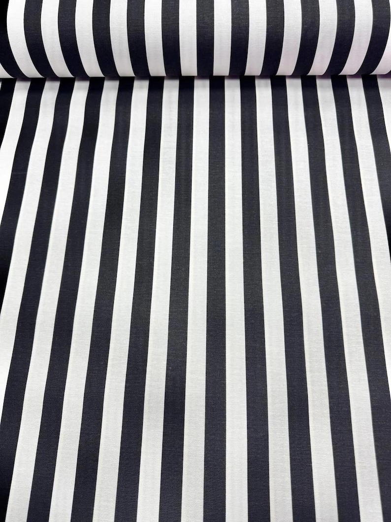 May include: Black and white striped fabric. The fabric is rolled up, showing the pattern of vertical stripes. The stripes alternate between black and white, creating a bold, graphic design. The fabric appears to be a medium weight material.
