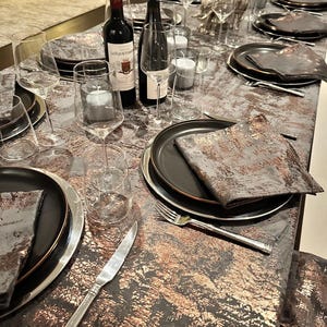 May include: A table setting with black and gold chargers, black napkins, and clear wine glasses. Two bottles of wine are on the table, one red and one white. The table is covered in a gray and gold tablecloth.