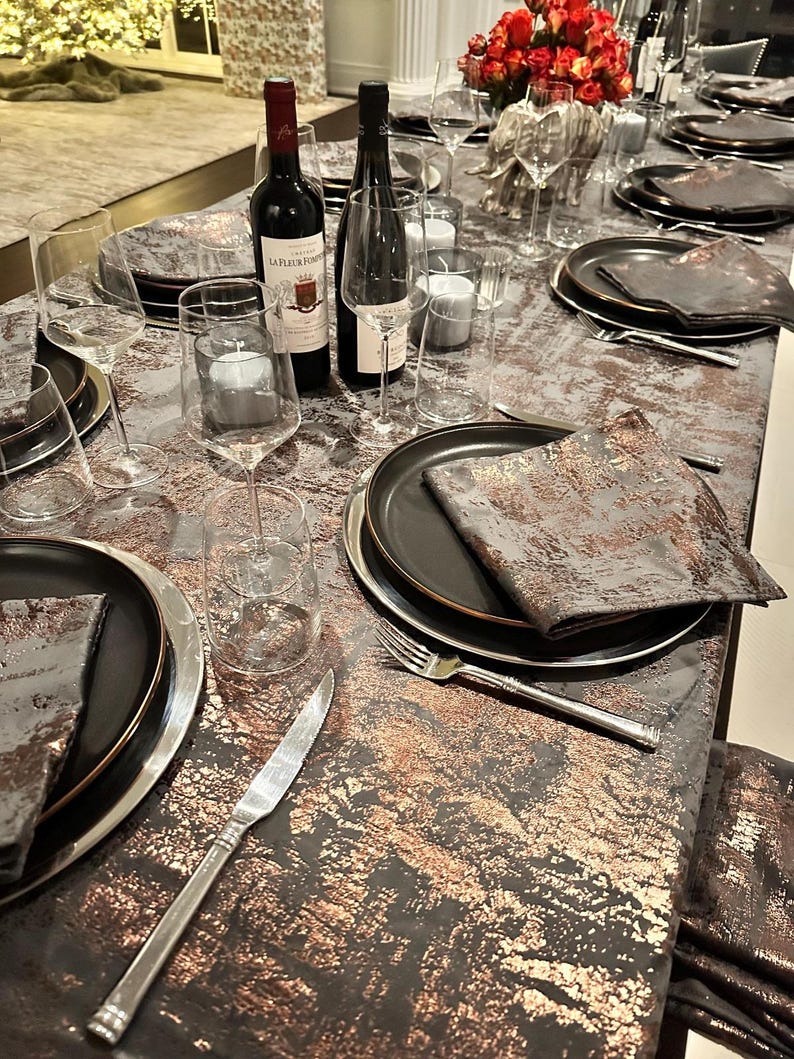 May include: A table setting with black and gold chargers, black napkins, and clear wine glasses. Two bottles of wine are on the table, one red and one white. The table is covered in a gray and gold tablecloth.