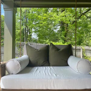 Puede incluir: Un columpio de cama de d&iacute;a blanco colgante con dos almohadas grandes verdes y dos almohadas blancas. El columpio est&aacute; colgado de un porche de madera con vista a &aacute;rboles verdes.