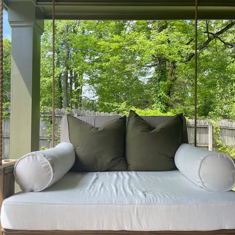 Puede incluir: Un columpio de cama de d&iacute;a blanco colgante con dos almohadas grandes verdes y dos almohadas blancas. El columpio est&aacute; colgado de un porche de madera con vista a &aacute;rboles verdes.