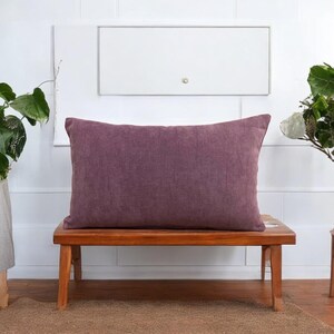 May include: A purple rectangular pillow sits on a wooden bench. The pillow is made of a textured fabric and has a simple design.