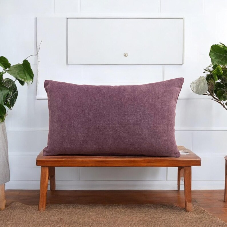 May include: A purple rectangular pillow sits on a wooden bench. The pillow is made of a textured fabric and has a simple design.