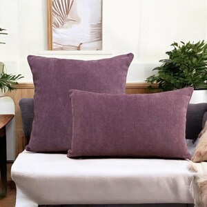 May include: Two purple decorative throw pillows on a white couch. The larger pillow is square and the smaller pillow is rectangular.