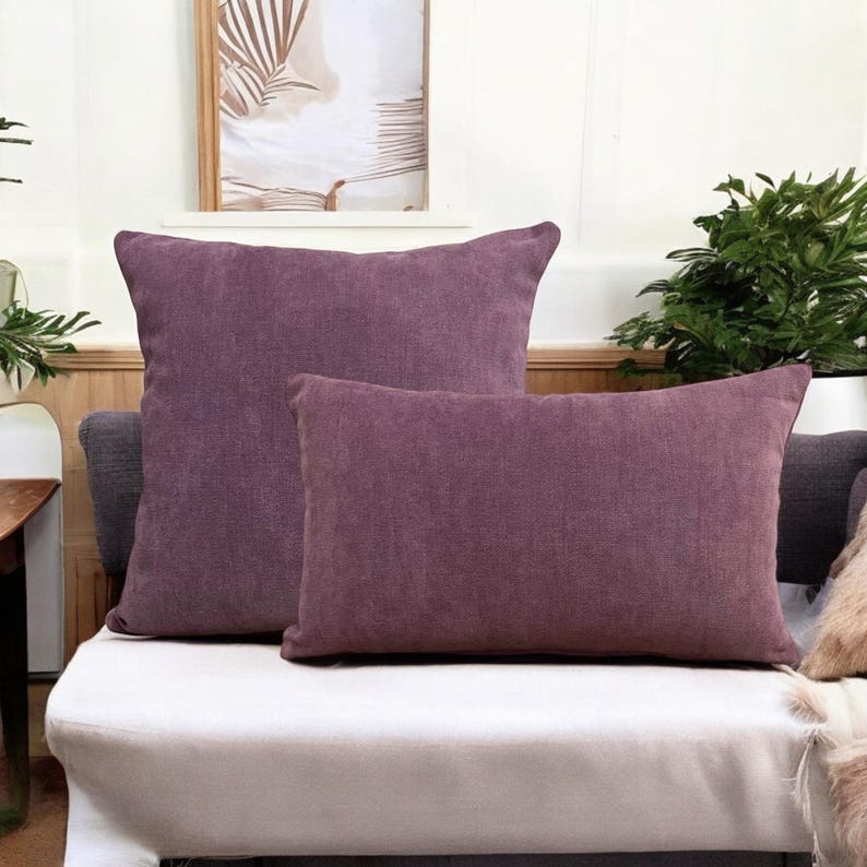 May include: Two purple decorative throw pillows on a white couch. The larger pillow is square and the smaller pillow is rectangular.