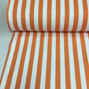 May include: A roll of fabric featuring vertical stripes in orange and white. The stripes are evenly spaced and run the length of the fabric. The fabric is tightly rolled, with the pattern visible on the surface.
