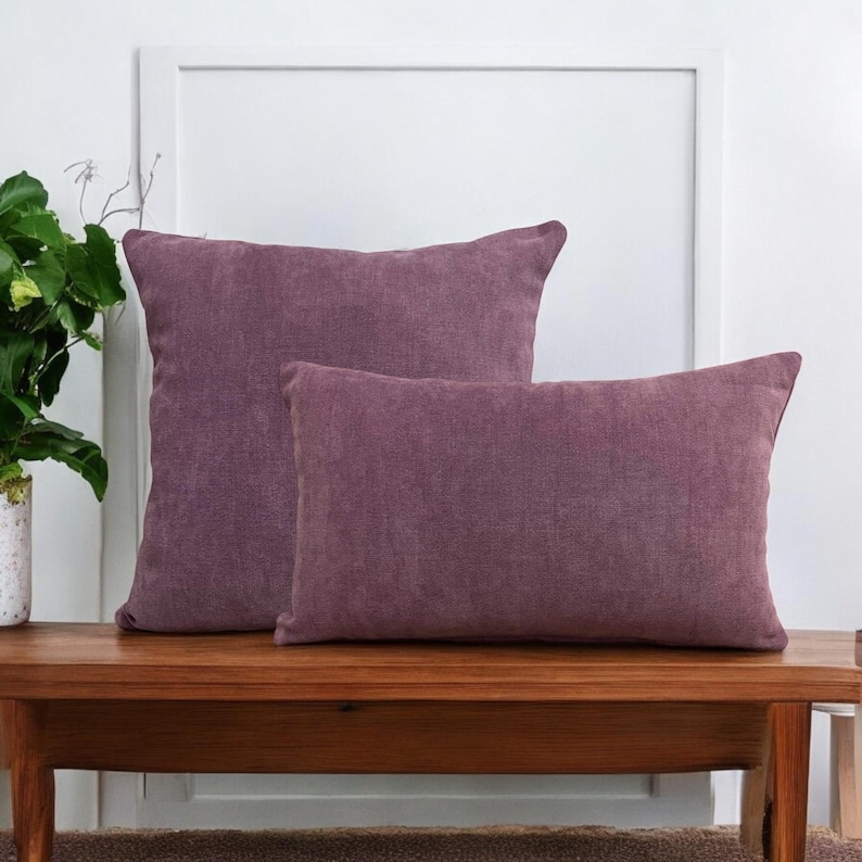 May include: Two decorative throw pillows in a dusty rose color. The pillows are made of a soft, textured fabric and are arranged on a wooden bench.