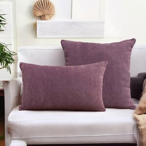 May include: Two purple decorative throw pillows on a white couch. The pillows are made of a soft, textured fabric.