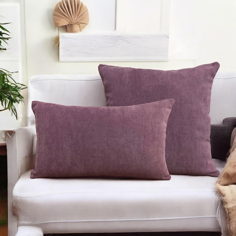 May include: Two purple decorative throw pillows on a white couch. The pillows are made of a soft, textured fabric.