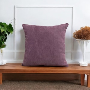 May include: A soft, purple throw pillow with a textured fabric. The pillow is sitting on a wooden bench.