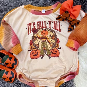 May include: A beige baby romper with long sleeves, featuring a pumpkin-themed graphic and the text "It's Fall Y'all." The sleeves and trim have an orange and pink tie-dye effect. A matching orange bow and pumpkin-faced shoes complete the fall ensemble.