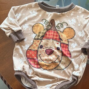 Pode incluir: Um macacão de manga comprida bege com um padrão floral branco e punhos e gola canelados castanhos. O centro apresenta um grande gráfico de um rosto de urso desenhado como uma abóbora de retalhos com um caule no topo.