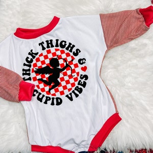 Może przedstawiać: Biały body z długim rękawem z czerwonymi i białymi paskami na rękawach. Z przodu znajduje się czarne i białe koło w kratkę z sylwetką Amora trzymającego strzałę i tekstem "Thick Thighs & Cupid Vibes".