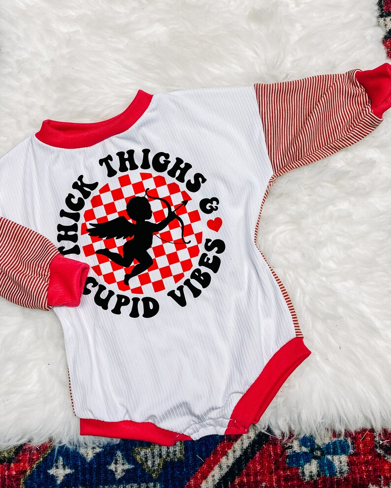 Może przedstawiać: Biały body z długim rękawem z czerwonymi i białymi paskami na rękawach. Z przodu znajduje się czarne i białe koło w kratkę z sylwetką Amora trzymającego strzałę i tekstem "Thick Thighs & Cupid Vibes".
