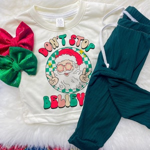 May include: White long-sleeved shirt with a green and red graphic of Santa Claus wearing sunglasses and the text "Don't Stop Believe".  The shirt is paired with dark green pants.