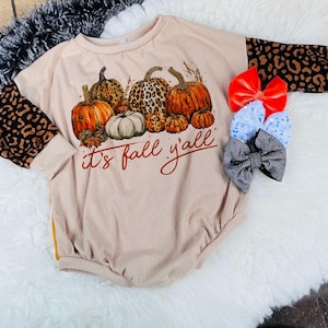 Pode incluir: Um body bege de manga comprida com um gráfico de abóboras e o texto "it's fall y'all". As mangas são feitas de tecido de estampa de leopardo marrom e preto.