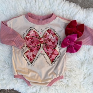 May include: A pink and white long-sleeved bodysuit with a large pink bow and glitter print. The text "You are loved you are enough You are worthy you are beautiful you are enough" is printed on the bodysuit.