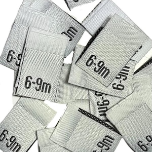 May include: A collection of small, rectangular woven labels in a light gray color. Each label has the text "6-9m" printed in black, indicating a size range. The labels are scattered, suggesting they are ready for use in clothing or textile projects.