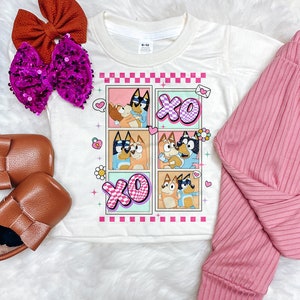 May include: White t-shirt with a graphic of cartoon dogs in a photo booth with the text "XO" on a pink and blue background. The shirt has a crew neck and short sleeves. The size tag says "6-12".