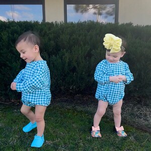 May include: Two toddlers wearing matching blue and white gingham rompers. The toddler on the right is wearing a yellow bow in their hair. Both toddlers are wearing blue shoes.