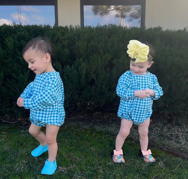 May include: Two toddlers wearing matching blue and white gingham rompers. The toddler on the right is wearing a yellow bow in their hair. Both toddlers are wearing blue shoes.