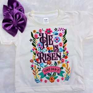 Könnte beinhalten: Weißes T-Shirt mit floralem Design und dem Text "He is Risen" mit dem Bibelvers "Lukas 24:6-7".