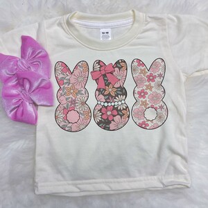 Könnte beinhalten: Ein weißes T-Shirt mit einem floralen Druck von drei Osterhasen. Die Hasen tragen rosa Schleifen und eine Perlenkette. Das Shirt hat ein Größenetikett mit der Aufschrift "12-18".