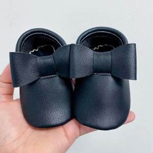 May include: A pair of black leather baby moccasins with a black bow on the front.