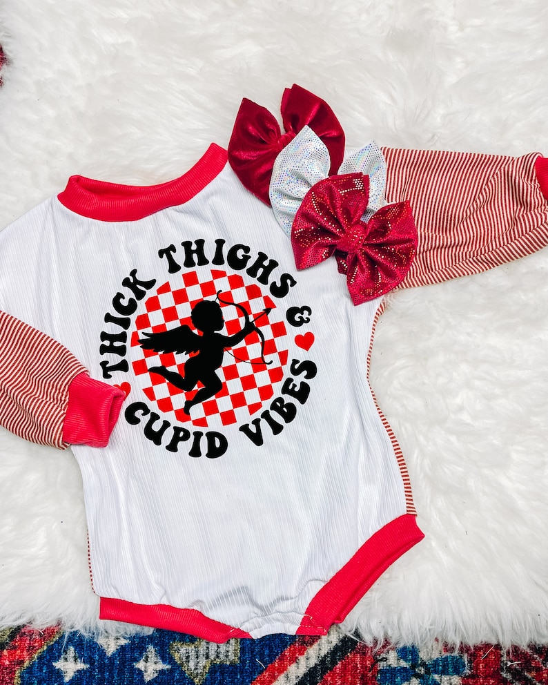 Może przedstawiać: Biały body z długim rękawem, z czerwonymi paskami na rękawach i czerwoną lam&oacute;wką wok&oacute;ł dekoltu. Body ma nadrukowany grafik przedstawiający amora na tle w kratkę i napis "Thick Thighs & Cupid Vibes".