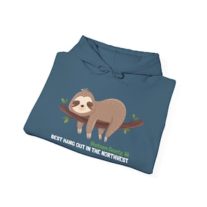 Könnte beinhalten: Ein marineblauer Kapuzenpullover mit einem Cartoon-Faultier-Design. Das Faultier ist braun und cremefarben und ruht auf einem braunen Ast mit grünen Blättern. Der Text auf dem Hoodie lautet "BEST HANG OUT IN THE NORTHWEST" und "Whatcom County, VA."