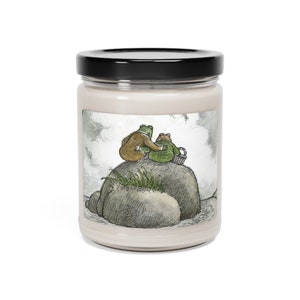 May include: A candle in a glass jar with a black lid. The label features an illustration of two frogs sitting on a rock, one in a brown coat and the other in a green jacket, with a basket. The candle has a cream-colored wax.