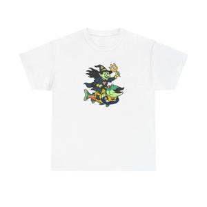 May include: White t-shirt featuring a cartoon illustration of a witch riding a fish. The witch is wearing a black hat and coat, holding a trident. The fish is yellow and blue. The design is centered on the front of the shirt.