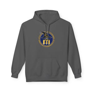 Peut inclure: Un sweat-shirt à capuche gris foncé avec une poche avant. Le sweat-shirt présente un graphisme circulaire avec un chapeau de sorcière, le texte "LET'S GO MARINERS" dans un anneau jaune et un emblème bleu et jaune.