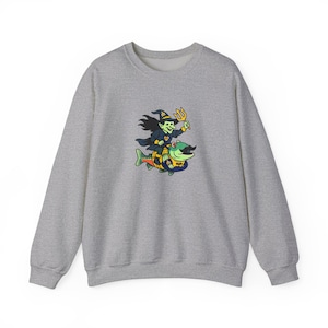 May include: A heather gray sweatshirt featuring a whimsical graphic. The graphic depicts a green witch riding a fish with a mustache, holding a trident. The witch wears a black hat and cape. The fish is yellow, blue, and green.