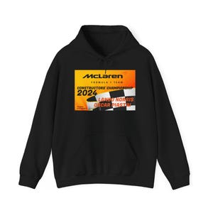 Może przedstawiać: Czarna bluza z kapturem z żółto-pomarańczową grafiką logo zespołu Formuły 1 McLaren. Grafika zawiera tekst "McLaren Formula 1 Team", "Constructors' Championship 2024", "Lando Norris" i "Oscar Piastri".