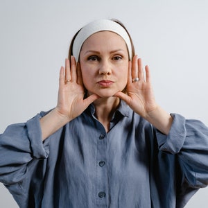 Puede incluir: Una mujer con una diadema blanca está realizando un ejercicio facial. Tiene las manos a ambos lados de la cara, con los dedos tocando la línea de la mandíbula. Lleva una camisa azul de botones.