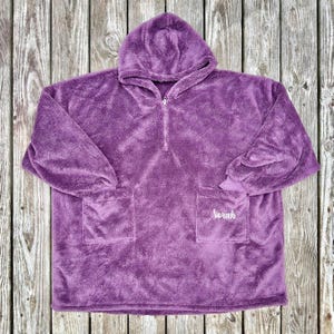 May include: A plush, purple hooded blanket with a half-zip closure and two front pockets. The name "Norah" is embroidered on one pocket. The blanket is made of a soft, fuzzy material.
