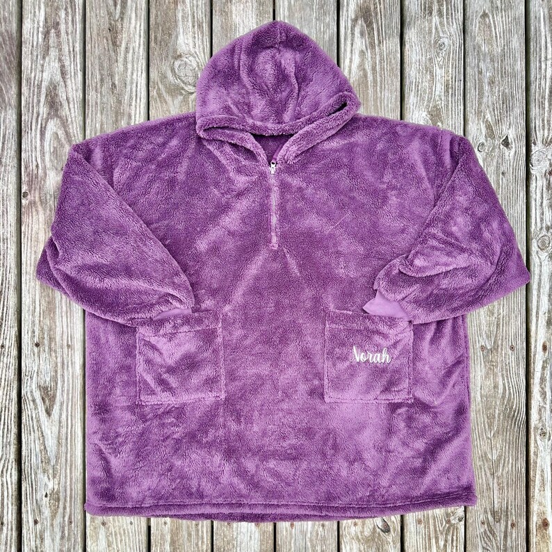 May include: A plush, purple hooded blanket with a half-zip closure and two front pockets. The name "Norah" is embroidered on one pocket. The blanket is made of a soft, fuzzy material.
