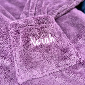 May include: Close-up of a plush, lavender-colored robe with a pocket. The pocket is embroidered with the name "Norah" in white script. The robe appears soft and cozy, perfect for lounging.