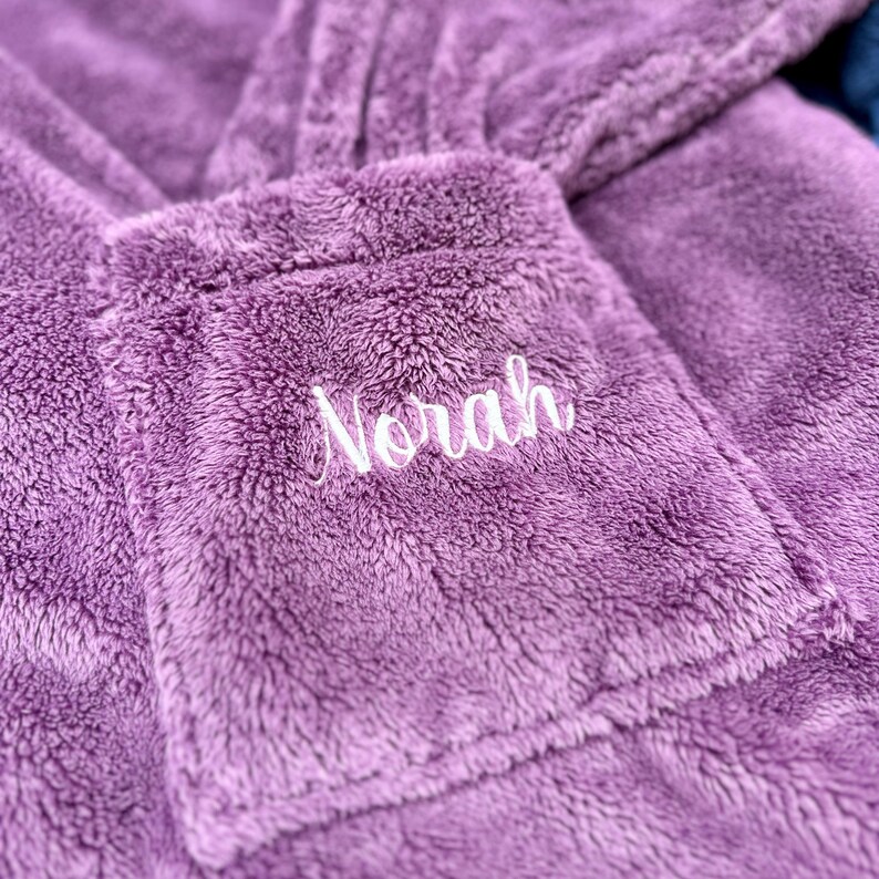 May include: Close-up of a plush, lavender-colored robe with a pocket. The pocket is embroidered with the name "Norah" in white script. The robe appears soft and cozy, perfect for lounging.