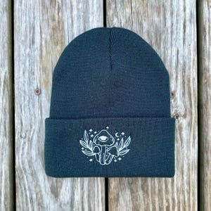 May include: Black beanie with a white embroidered design of a mushroom with an eye, surrounded by leaves and stars.