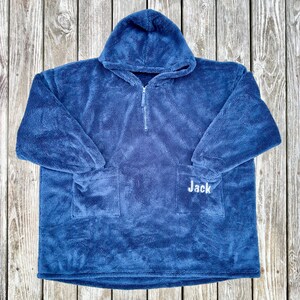 May include: A blue hooded blanket with a front pocket and a half-zip closure. The name "Jack" is embroidered in white on the front pocket. The blanket is made of a soft, plush material.