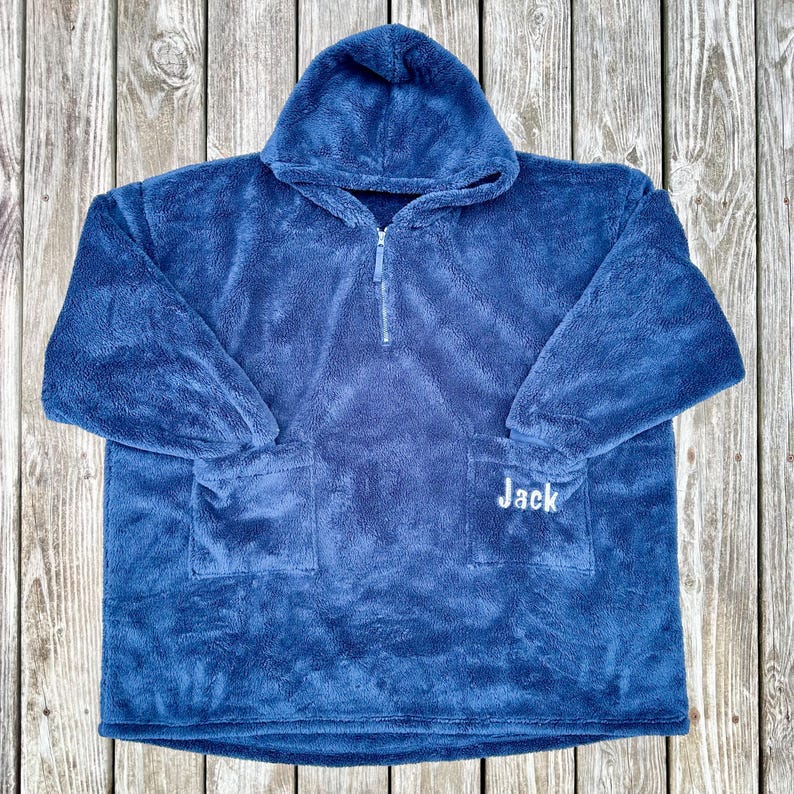 May include: A blue hooded blanket with a front pocket and a half-zip closure. The name "Jack" is embroidered in white on the front pocket. The blanket is made of a soft, plush material.