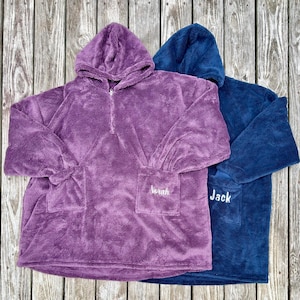 May include: Two plush hooded sweatshirts in purple and navy blue. The purple sweatshirt has the name "Sarah" embroidered on the pocket. The navy blue sweatshirt has the name "Jack" embroidered on the pocket. Both sweatshirts have a front pocket and a zipper at the neck.