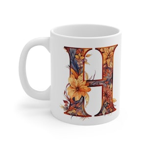 Puede incluir: Taza de cerámica blanca con la letra "H" en un diseño floral. La letra está llena de flores y follaje naranja, amarillo y marrón. La taza tiene un asa estándar y un acabado brillante, adecuada para bebidas calientes.