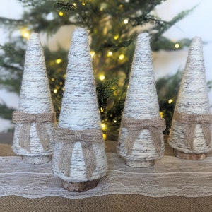 May include: Four white and brown yarn wrapped cone shaped Christmas trees with burlap bows. The trees are sitting on a white lace table runner.