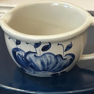 May include: Cream-colored ceramic bowl with a handle and spout. The bowl is decorated with blue floral designs. The handle is curved, and the spout is on the opposite side. The bowl is sitting on a blue surface.
