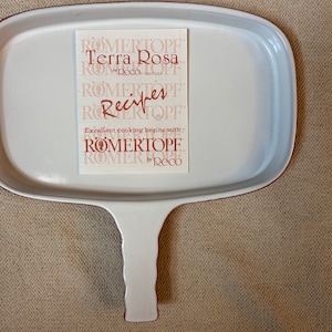 May include: White ceramic baking dish with a brown rim and a long handle. The dish is sitting on a brown surface. A white card with the text "Römertopf Terra Rosa by Reco" is in the dish.