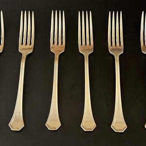 May include: Six silver forks with ornate handles. The forks are arranged in a row on a black surface.